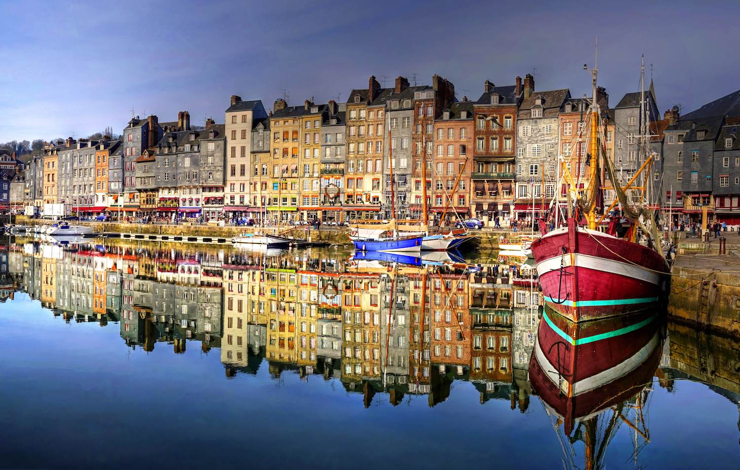 Panoramic view of Honfleur harbor in Normandy, France, featuring colorful historic buildings, traditional fishing boats, and the picturesque waterfront at sunset.